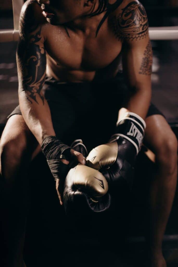 Microbursts for longevity and telomere health - A muscular boxer with tattoos sitting shirtless in a gym holding boxing gloves.